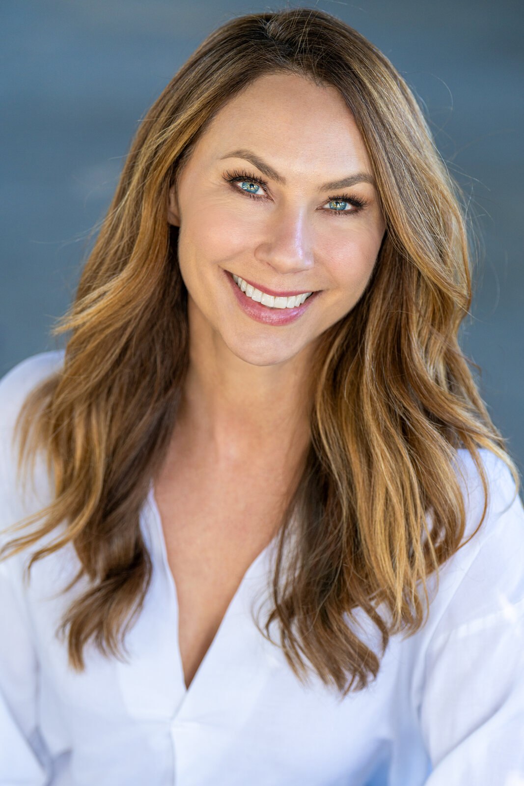 Smiling person with long hair in a white shirt against a blue-gray background.